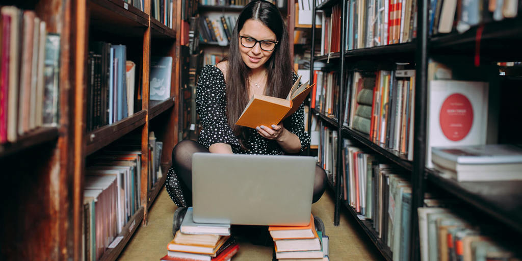 woman reading book and working on laptop in between bookcases in library